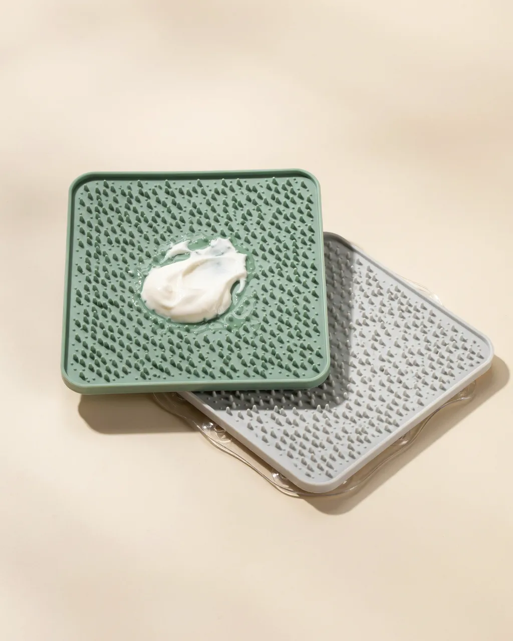 Tapis de léchage vert sauge garni de yaourt posé sur un tapis gris, avec les ventouses transparentes au dos, sur fond crème